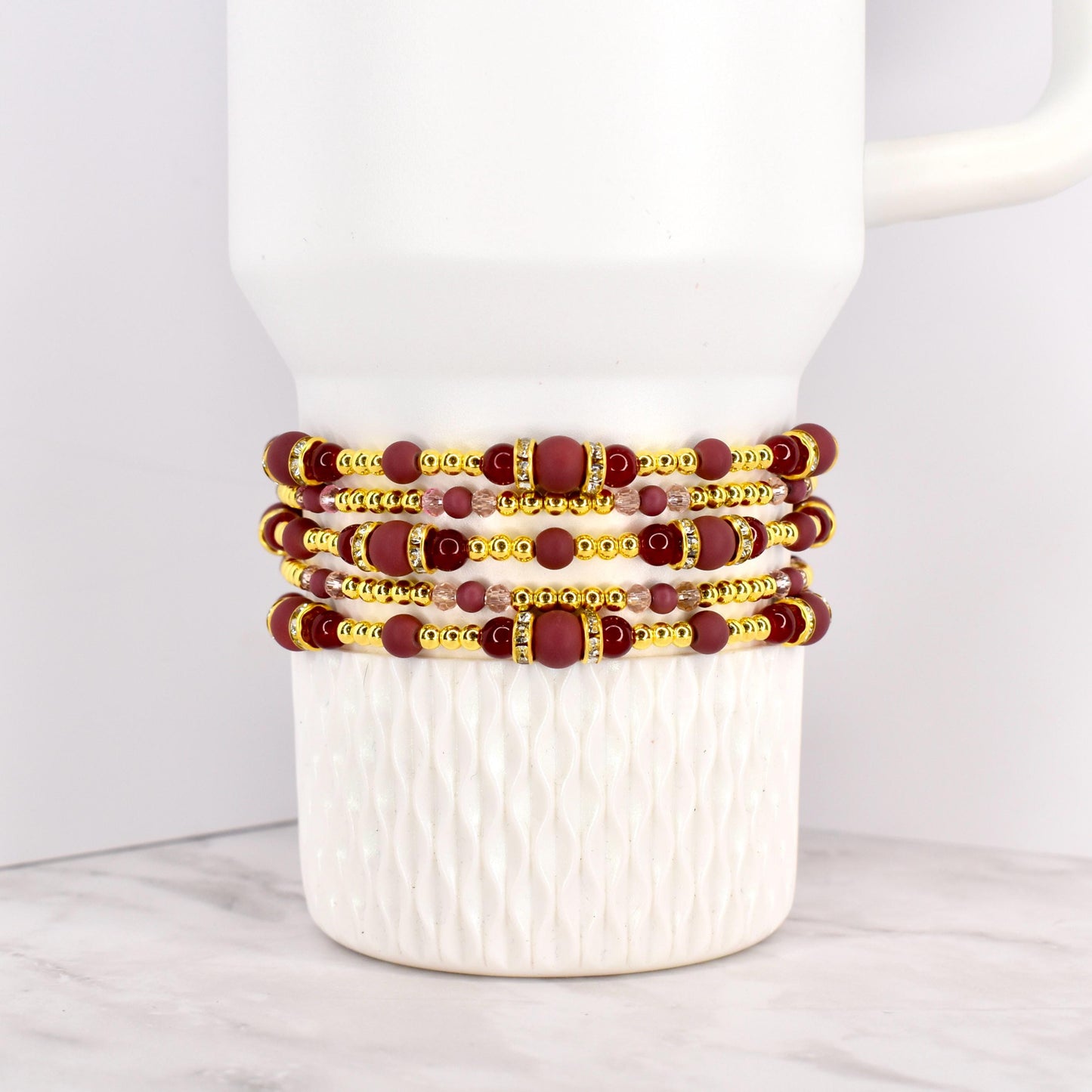 Four beaded bracelets in shades of burgundy, gold, and clear crystals are stacked on a white mug. The bracelets feature a mix of round and rectangular beads, with gold accents. The bracelets are likely for wrist wear.