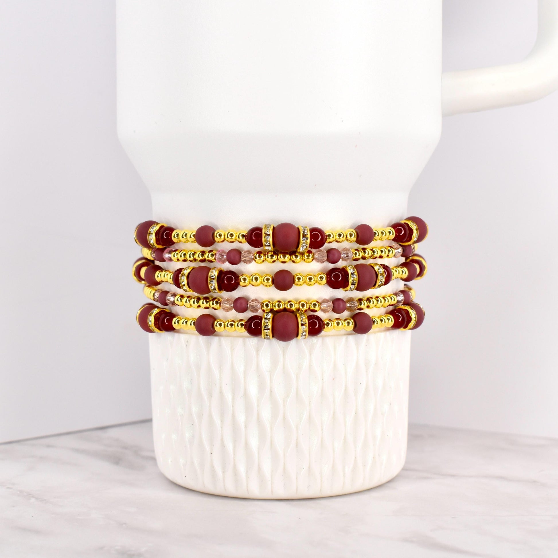 Four beaded bracelets in shades of burgundy, gold, and clear crystals are stacked on a white mug. The bracelets feature a mix of round and rectangular beads, with gold accents. The bracelets are likely for wrist wear.