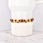 Two beaded bracelets. The top bracelet features maroon beads and gold accents, with a rectangular maroon center piece. The bottom bracelet has smaller pink beads and gold accents. The bracelets are stacked on a white ceramic container.