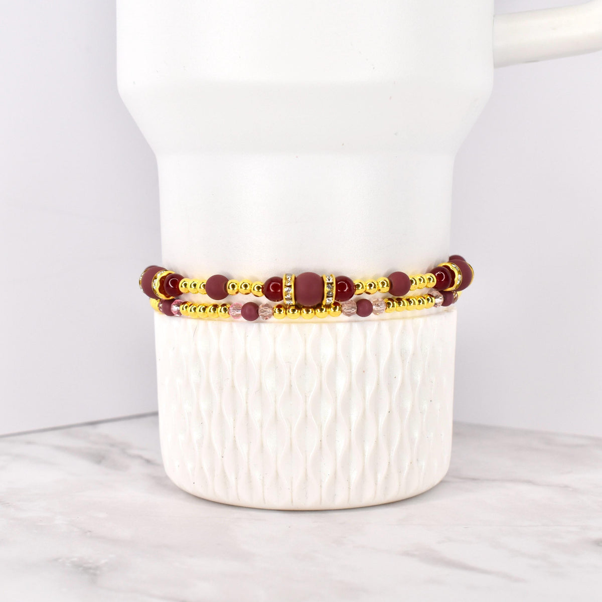 Two beaded bracelets. The top bracelet features maroon beads and gold accents, with a rectangular maroon center piece. The bottom bracelet has smaller pink beads and gold accents. The bracelets are stacked on a white ceramic container.