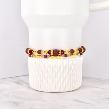 Two beaded bracelets. The top bracelet features maroon beads and gold accents, with a rectangular maroon center piece. The bottom bracelet has smaller pink beads and gold accents. The bracelets are stacked on a white ceramic container.