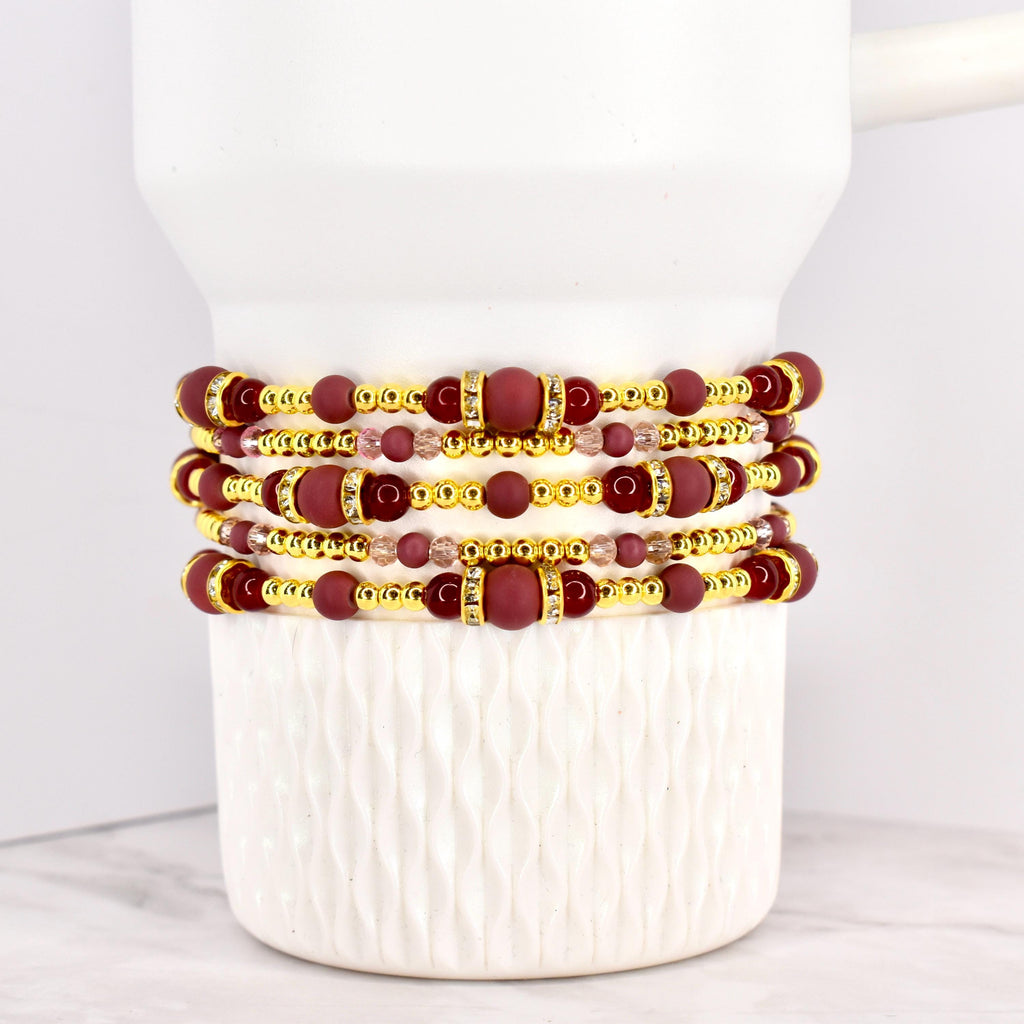 Four beaded bracelets in shades of burgundy, gold, and clear crystals are stacked on a white ceramic mug. The bracelets feature a mix of round and cylindrical beads, with gold accents.