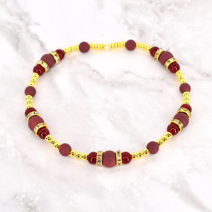 A beaded bracelet featuring a combination of burgundy and gold-colored beads. The bracelet has a repeating pattern of burgundy beads and gold-colored accents, with some sections adorned with small, sparkling embellishments. The bracelet is displayed on a white marble surface.