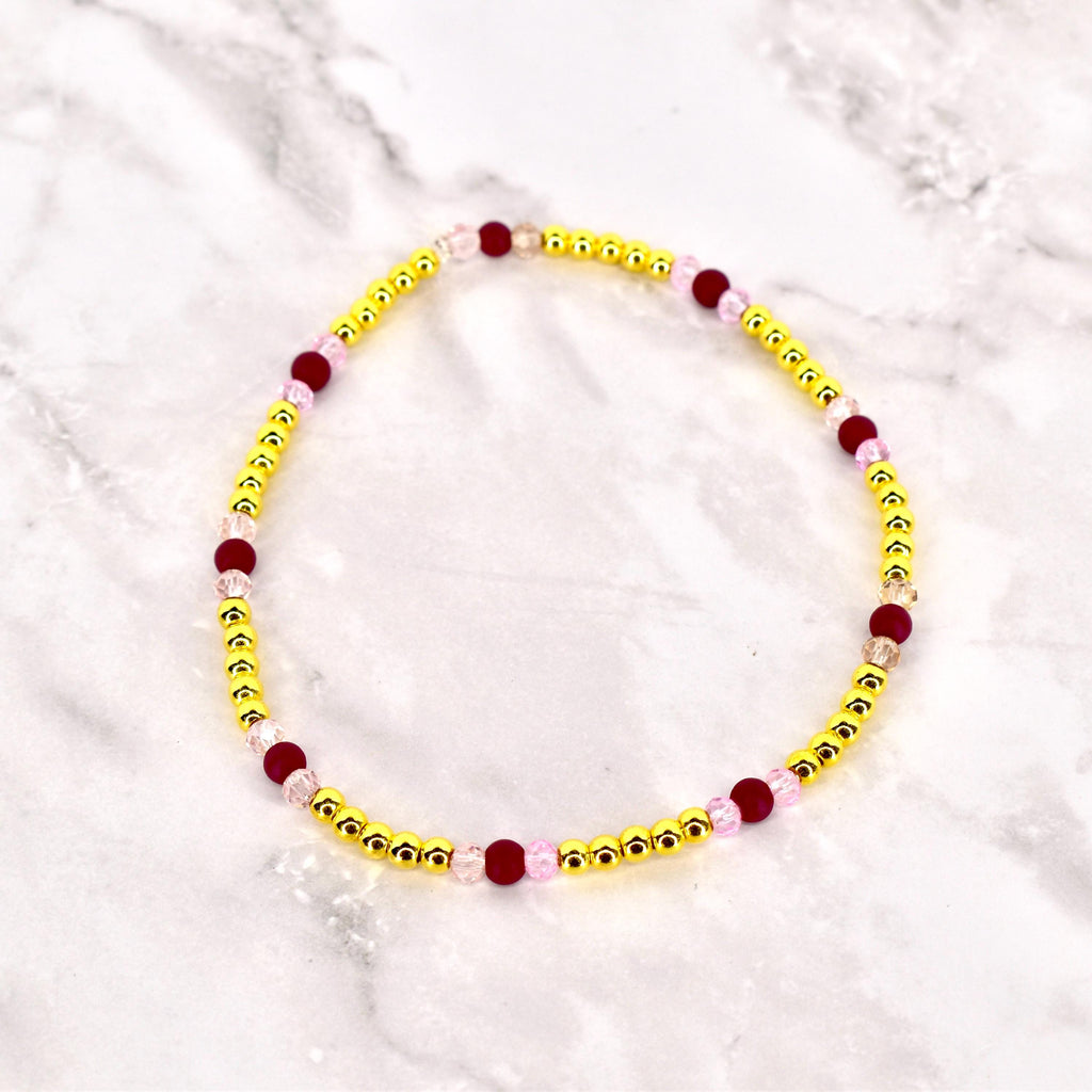 A beaded bracelet featuring small, gold-colored beads interspersed with larger, dark red and light pink beads. The bracelet is circular and likely designed to be worn around the wrist. The beads are strung together, creating a delicate and colorful accessory.