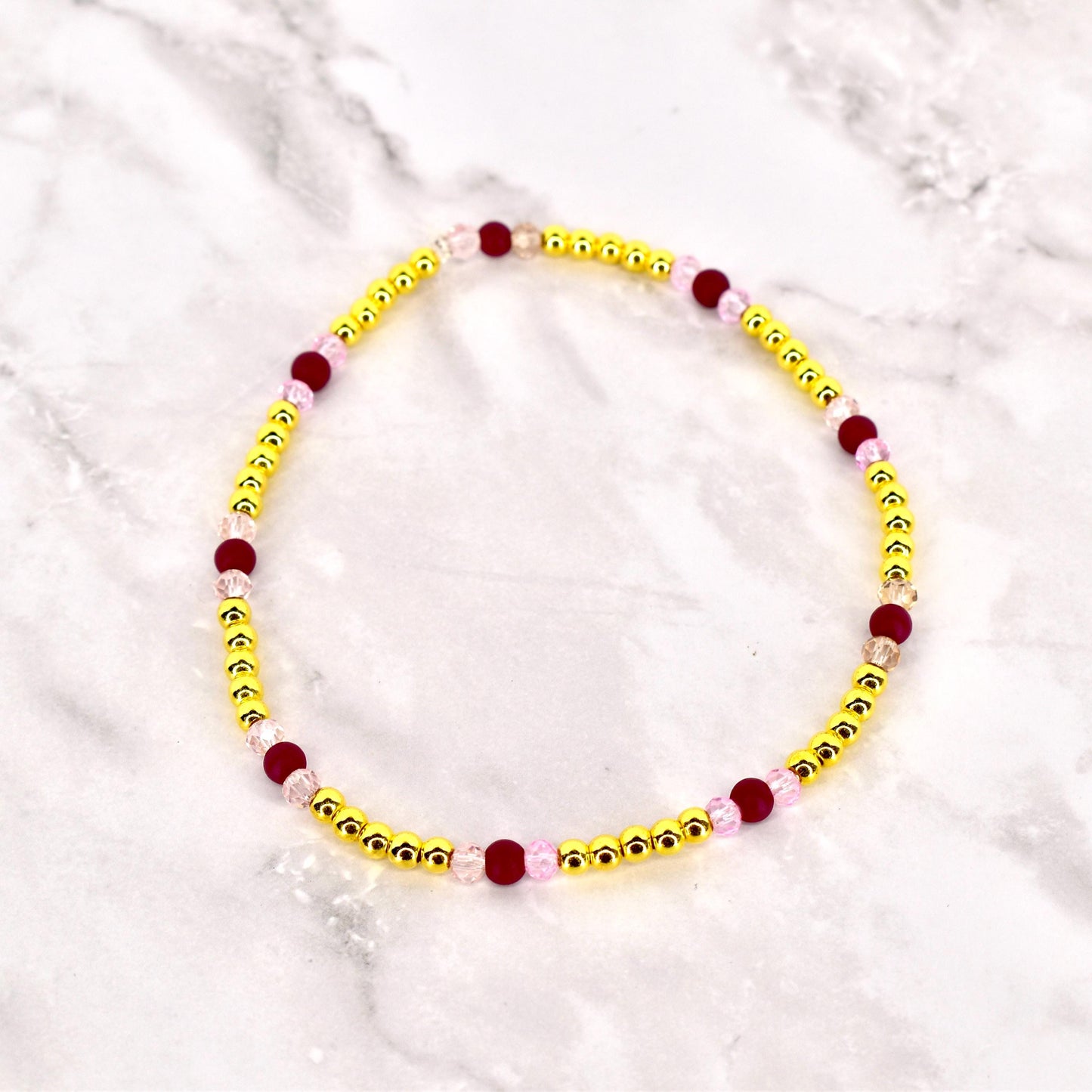 A beaded bracelet featuring small, gold-colored beads interspersed with larger, dark red and light pink beads. The bracelet is circular and likely designed to be worn around the wrist. The beads are strung together, creating a delicate and colorful accessory.