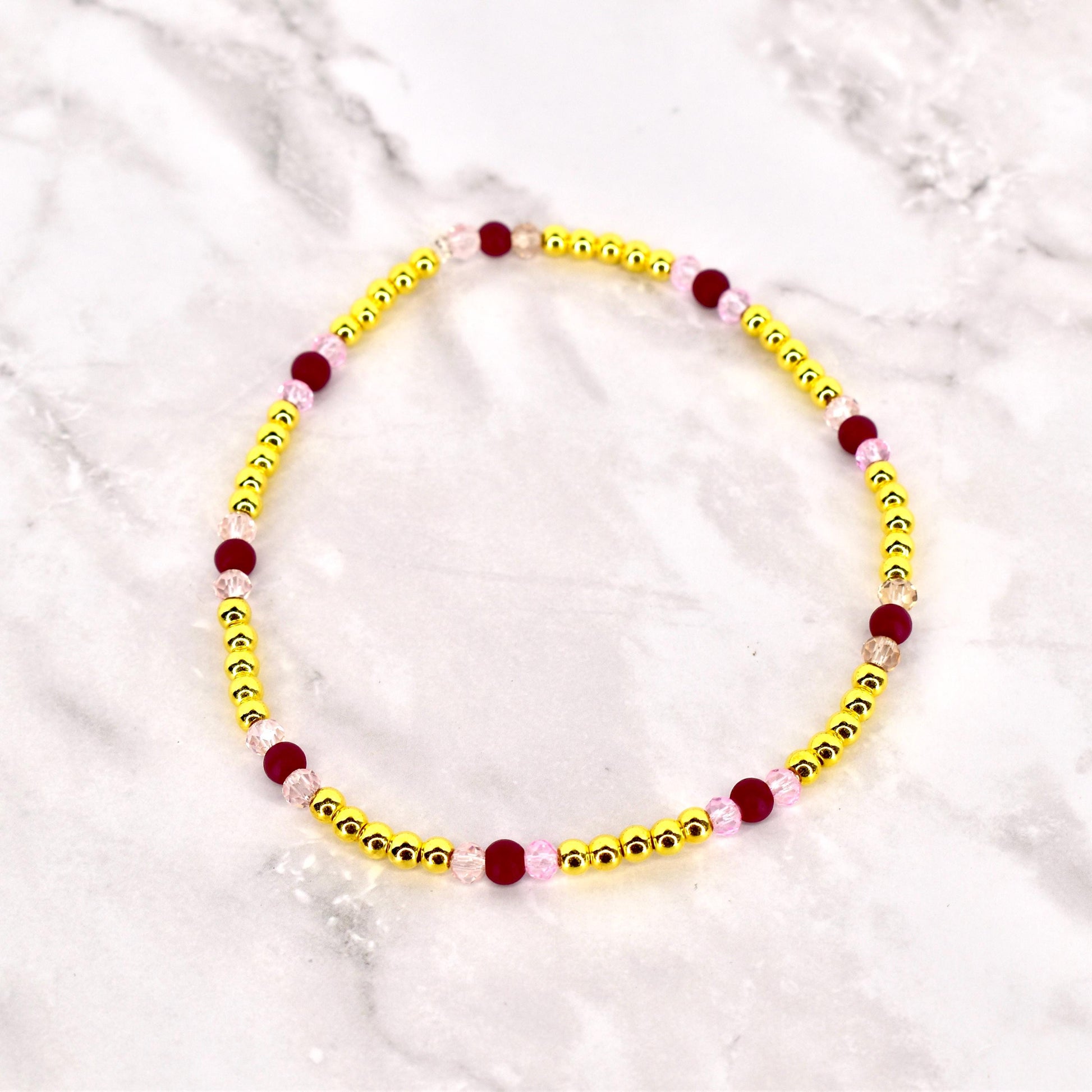 A beaded bracelet featuring small, gold-colored beads interspersed with larger, dark red and light pink beads. The bracelet is circular and likely designed to be worn around the wrist. The beads are strung together, creating a delicate and colorful accessory.