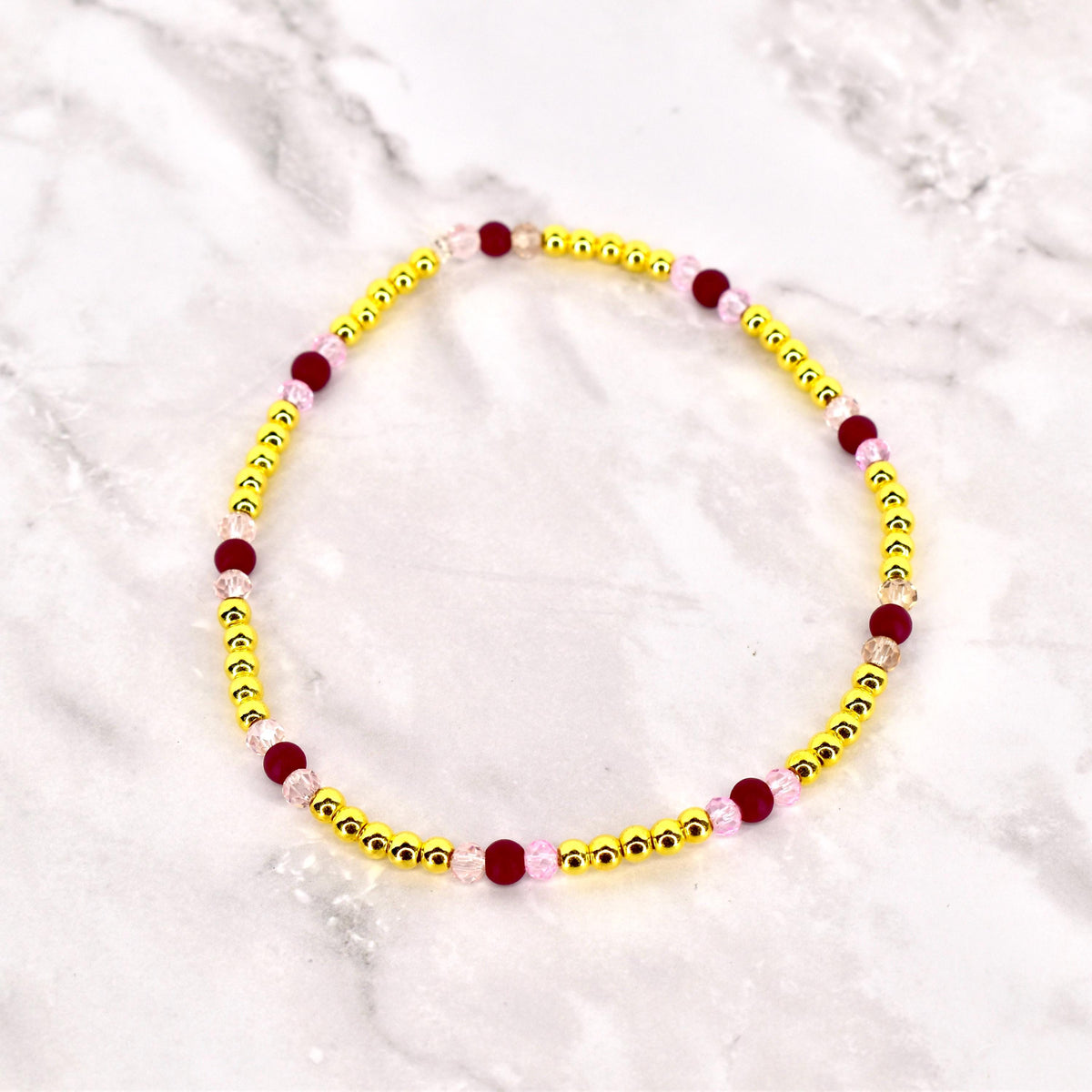 A beaded bracelet featuring small, gold-colored beads interspersed with larger, dark red and light pink beads. The bracelet is circular and likely designed to be worn around the wrist. The beads are strung together, creating a delicate and colorful accessory.
