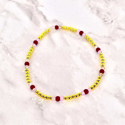 A beaded bracelet featuring small, gold-colored beads interspersed with larger, dark red and light pink beads. The bracelet is circular and likely designed to be worn around the wrist. The beads are strung together, creating a delicate and colorful accessory.