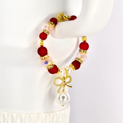 A red and gold beaded Stanley handle bracelet with a gold bow and pearl pendant. The bracelet features red, clear, and pink beads, with gold accents. The pendant includes a gold bow and a large white pearl.