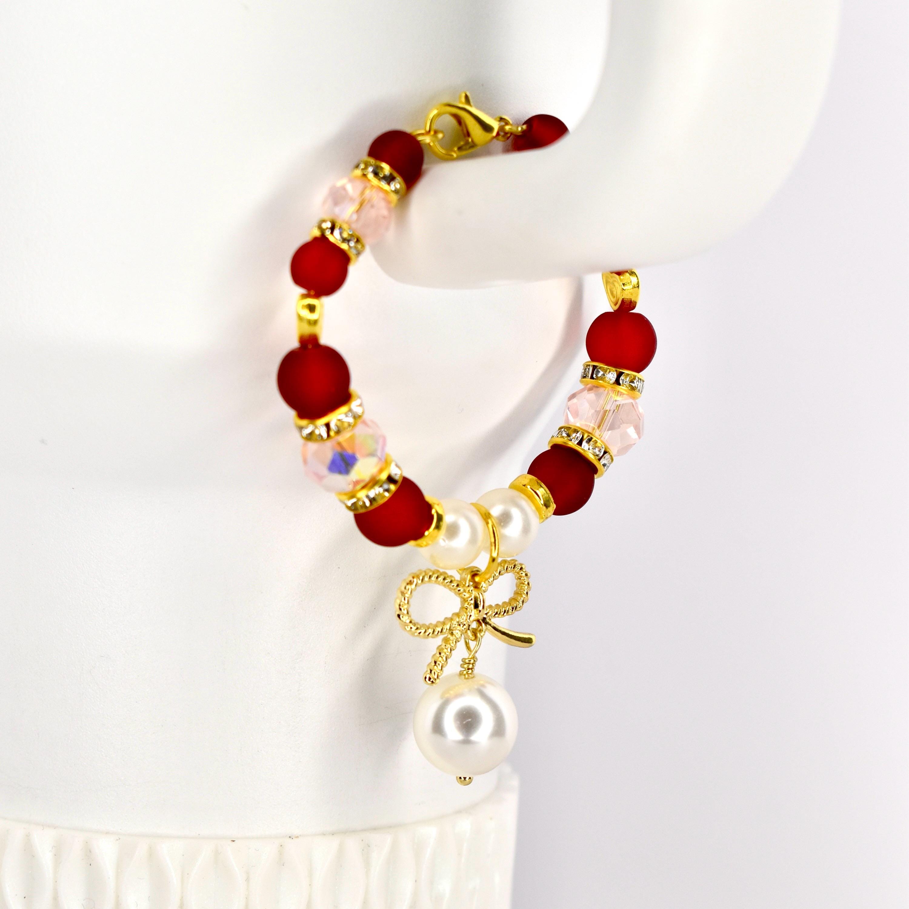 A red and gold beaded Stanley handle bracelet with a gold bow and pearl pendant. The bracelet features red, clear, and pink beads, with gold accents. The pendant includes a gold bow and a large white pearl.