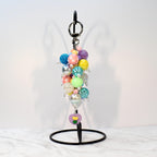 A colorful beaded keychain with a variety of pastel and iridescent beads. The keychain includes a silver keyring and a charm with a purple heart and yellow flower design. The beads are in shades of pink, blue, yellow, and green.