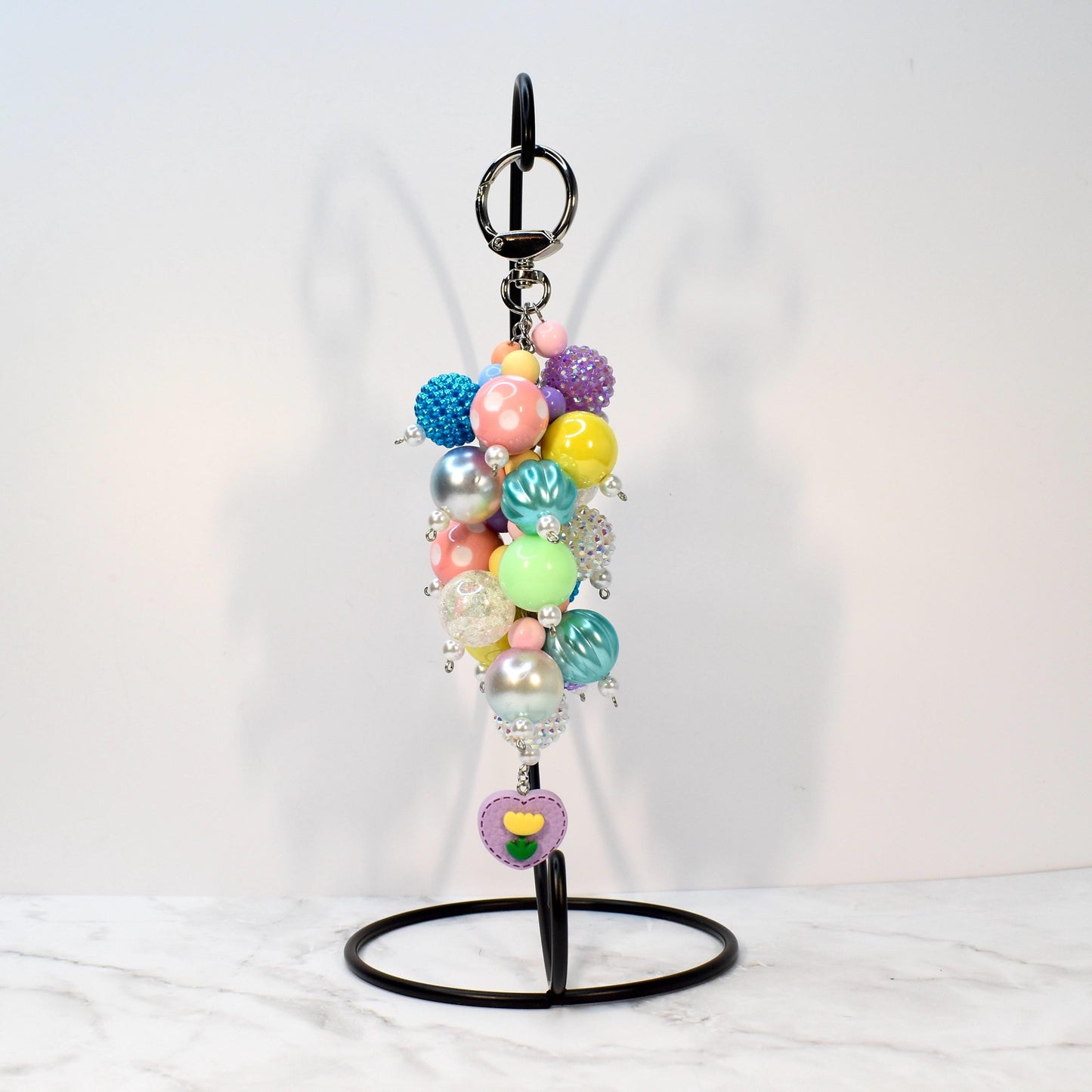 A colorful beaded keychain with a variety of pastel and iridescent beads. The keychain includes a silver keyring and a charm with a purple heart and yellow flower design. The beads are in shades of pink, blue, yellow, and green.