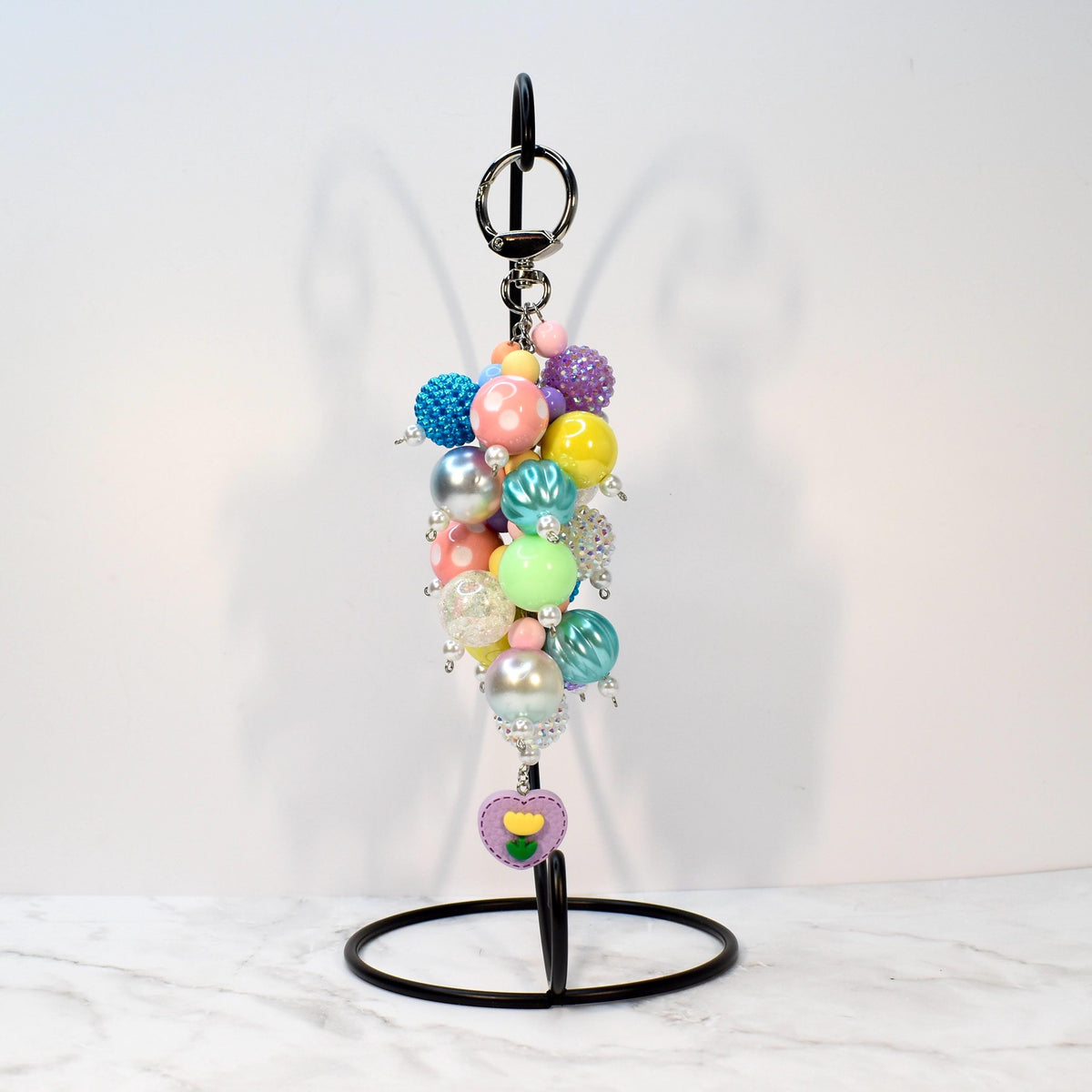 A colorful beaded keychain with a variety of pastel and iridescent beads. The keychain includes a silver keyring and a charm with a purple heart and yellow flower design. The beads are in shades of pink, blue, yellow, and green.
