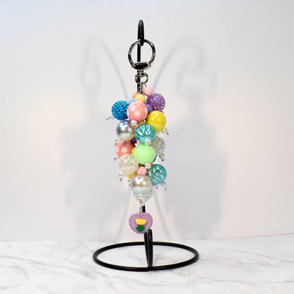 A colorful beaded keychain with a variety of pastel and iridescent beads. The keychain includes a silver keyring and a charm with a purple heart and yellow flower design. The beads are in shades of pink, blue, yellow, and green.