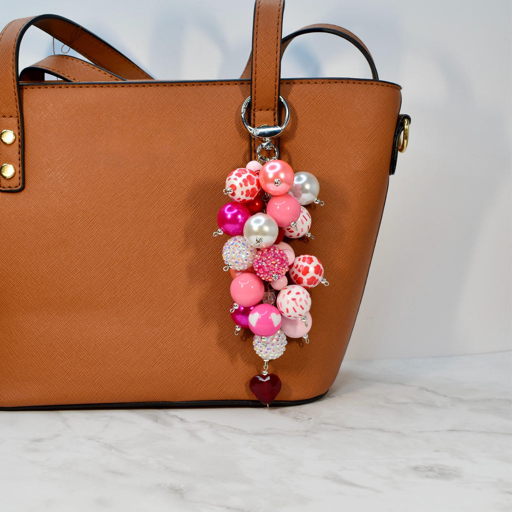 A cluster of pink and white beads with a silver keychain attached to a brown handbag. The beads vary in size and pattern, including solid pink, white with red polka dots, and heart-shaped designs. A small, dark red heart charm hangs at the bottom.