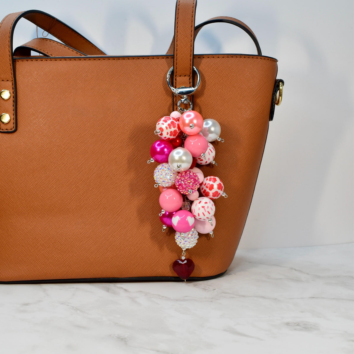 A cluster of pink and white beads with a silver keychain attached to a brown handbag. The beads vary in size and pattern, including solid pink, white with red polka dots, and heart-shaped designs. A small, dark red heart charm hangs at the bottom.