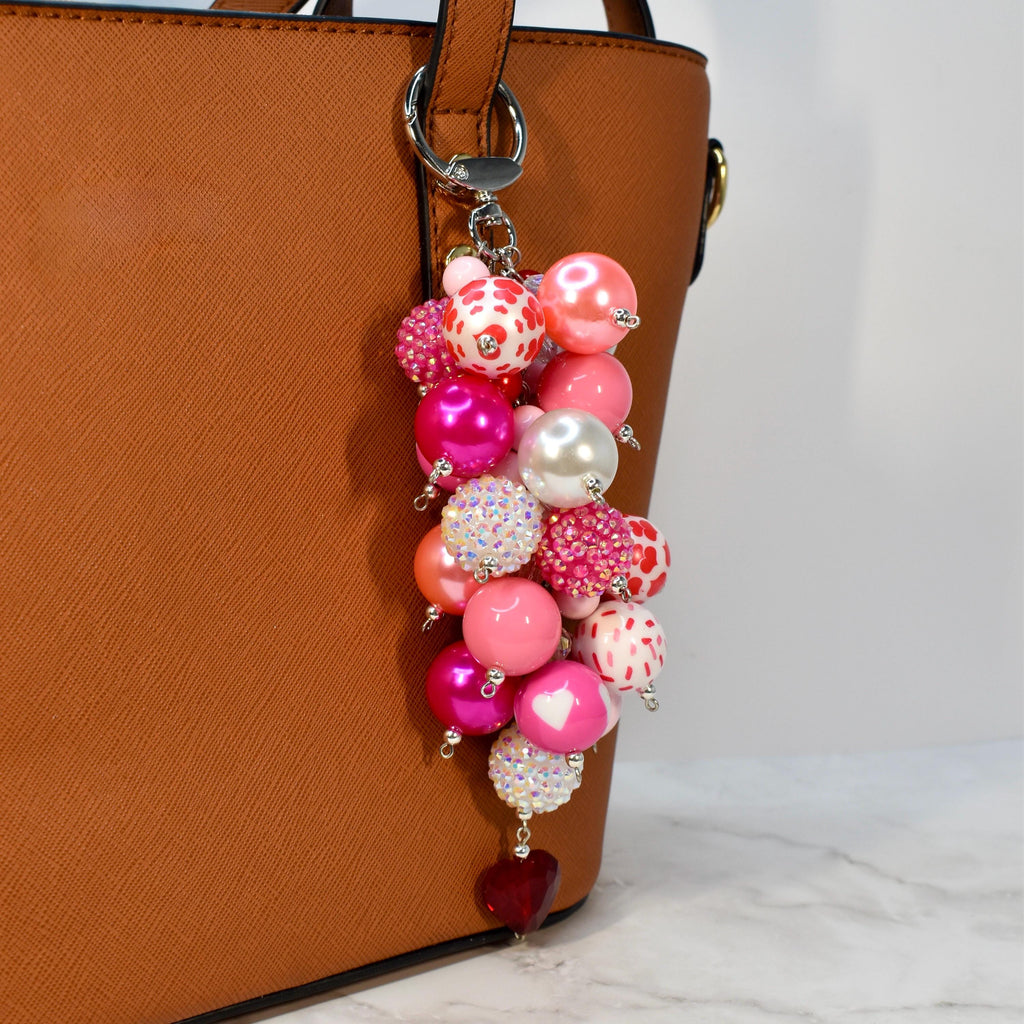 A beaded keychain with a cluster of pink, white, and red beads. The keychain features a variety of bead sizes and textures, including pearls, rhinestones, and heart-shaped beads. A red heart-shaped charm hangs at the bottom.