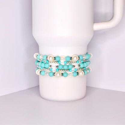 A stack of five bracelets made of turquoise blue and white beads. The bracelets are stacked on top of each other and are on a white surface.