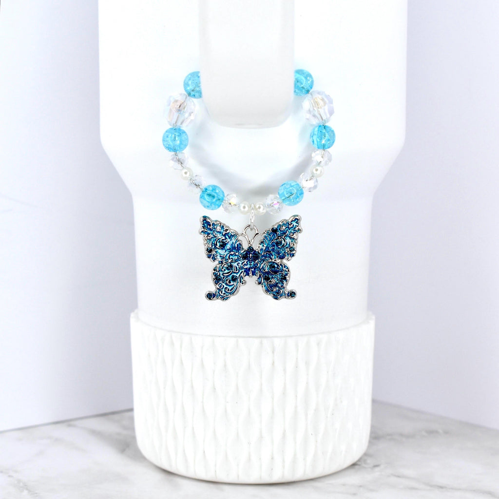 A blue butterfly pendant with intricate detailing hangs from a beaded necklace. The necklace features clear and light blue beads. The butterfly is a deep blue color and is displayed on a white background.
