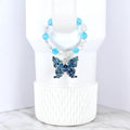 A blue butterfly pendant with intricate detailing hangs from a beaded necklace. The necklace features clear and light blue beads. The butterfly is a deep blue color and is displayed on a white background.