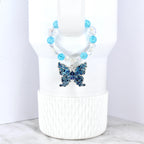 A blue butterfly pendant with intricate detailing hangs from a beaded necklace. The necklace features clear and light blue beads. The butterfly is a deep blue color and is displayed on a white background.