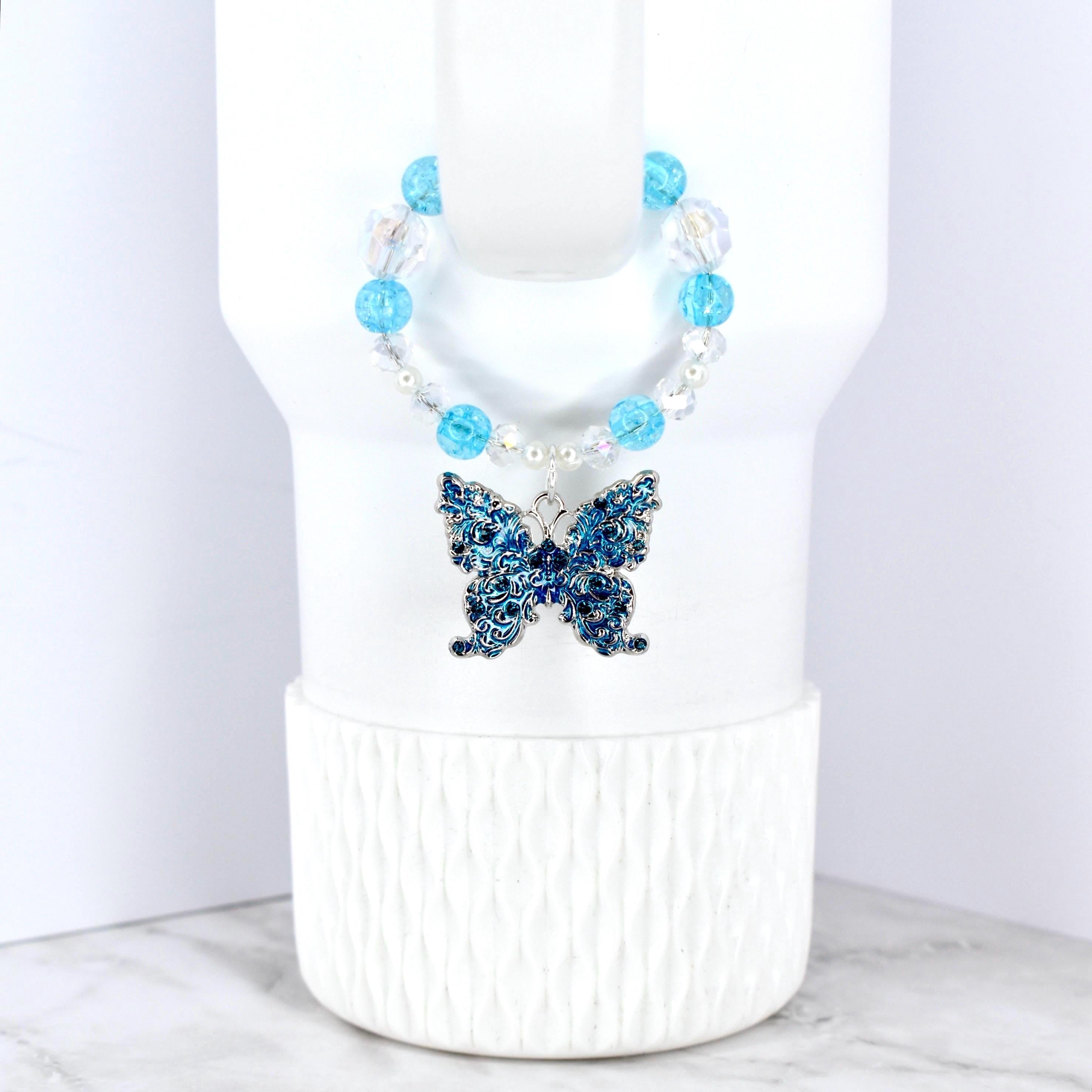 A blue butterfly pendant with intricate detailing hangs from a beaded necklace. The necklace features clear and light blue beads. The butterfly is a deep blue color and is displayed on a white background.