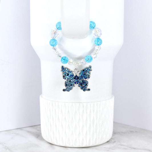 A blue butterfly pendant with intricate detailing hangs from a beaded necklace. The necklace features clear and light blue beads. The butterfly is a deep blue color and is displayed on a white background.