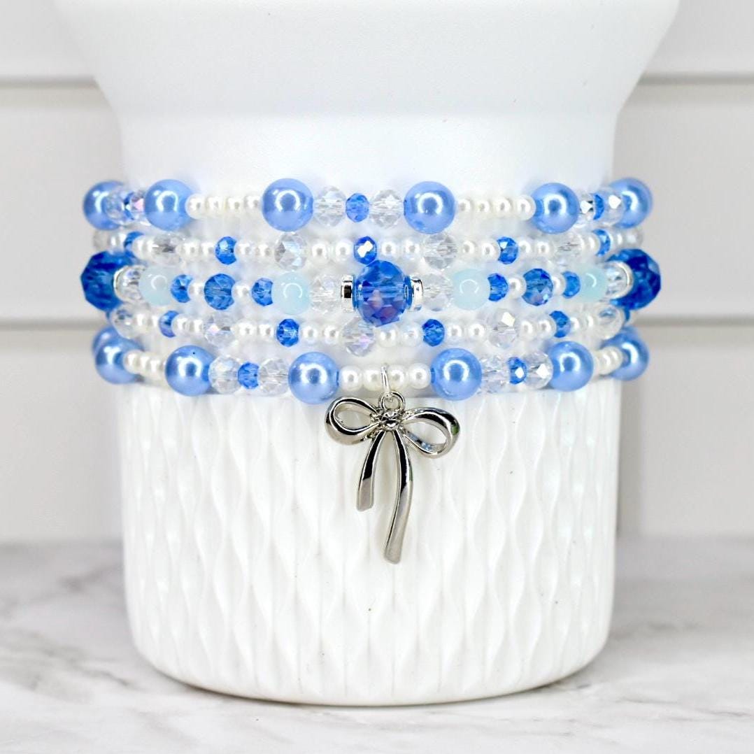 A multi-strand bracelet with blue, white, and clear beads. The bracelet has a silver bow charm hanging from it. The beads are various sizes and shades of blue, with some clear crystal beads interspersed.