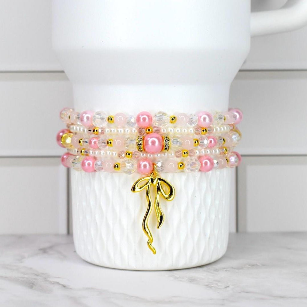 Stack of pink and clear beaded bracelets with gold accents and a gold charm. The bracelets are displayed on a white, textured surface. The charm is a gold ribbon design.