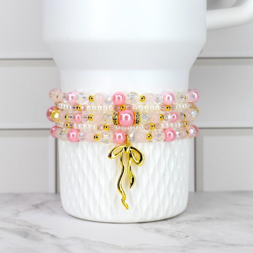 Stack of pink and clear beaded bracelets with gold accents and a gold charm. The bracelets are displayed on a white, textured surface. The charm is a gold ribbon design.