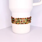 A collection of beaded bracelets in shades of green, gold, and brown are wrapped around a white mug. The bracelets appear to be made of small, colorful beads, creating a layered effect. The bracelets are a mix of matte and shiny beads.