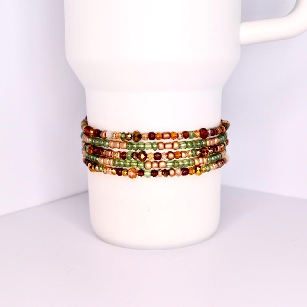 A collection of beaded bracelets in shades of green, gold, and brown are wrapped around a white mug. The bracelets appear to be made of small, colorful beads, creating a layered effect. The bracelets are a mix of matte and shiny beads.