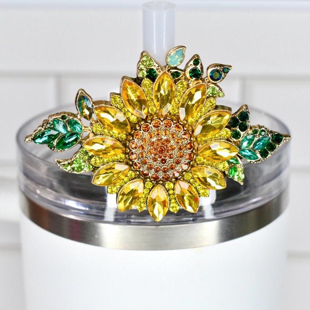 A decorative sunflower straw topper with yellow, orange, and green jewel-like accents. The sunflower is attached to a clear lid on a white tumbler. The topper adds a touch of sparkle and charm.