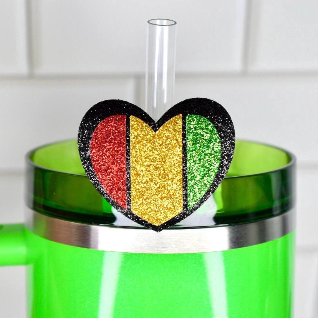 A green tumbler with a heart-shaped straw topper. The topper is divided into red, gold, and green sections with black glitter edging. A clear straw extends from the topper.