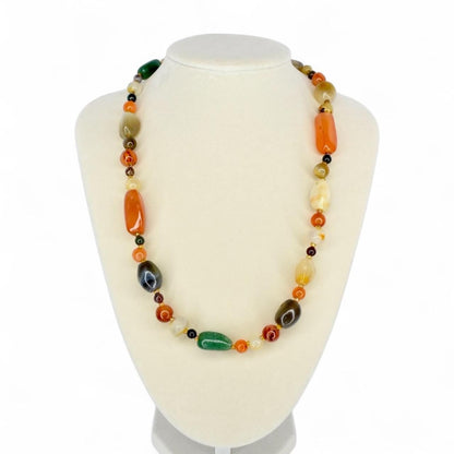 A colorful necklace with a variety of polished stone beads in shades of orange, green, brown, and yellow. The necklace is displayed on a white mannequin bust, showcasing its design and color variations.