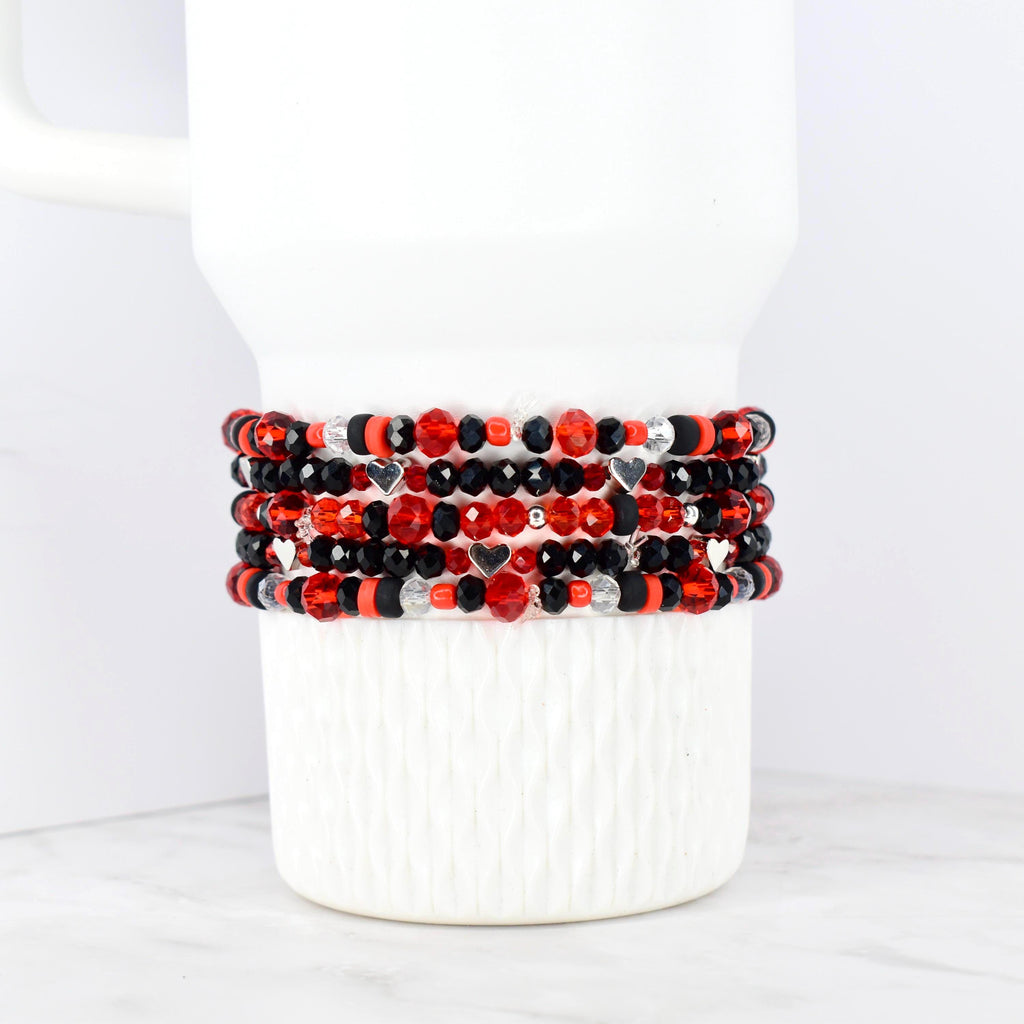 Stack of beaded bracelets in red, black, and silver. The bracelets feature a variety of beads, including faceted and round beads, with small silver heart charms. The bracelets are displayed on a white, textured surface.