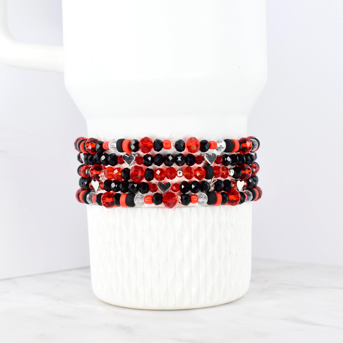 Stack of beaded bracelets in red, black, and silver. The bracelets feature a variety of beads, including faceted and round beads, with small silver heart charms. The bracelets are displayed on a white, textured surface.