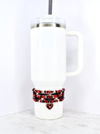 White insulated tumbler with a handle and a clear lid. A beaded wrap in red, black, and silver beads encircles the base, with a heart charm. The tumbler has a black straw.