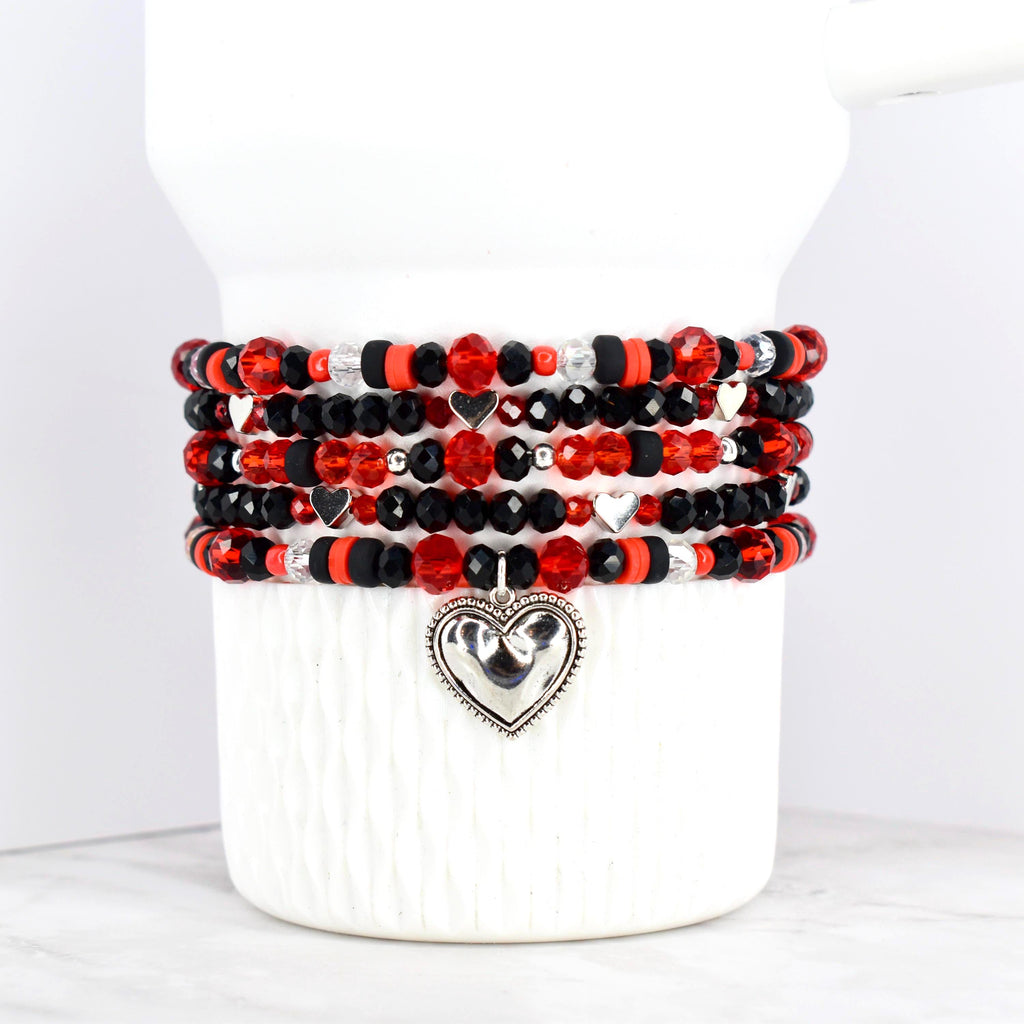 Stack of beaded bracelets in red, black, and silver. The bracelets feature a variety of beads, including faceted and round beads. Each bracelet has a silver heart charm. The bracelets are displayed on a white ceramic container.