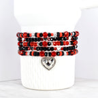 Stack of beaded bracelets in red, black, and silver. The bracelets feature a variety of beads, including faceted and round beads. Each bracelet has a silver heart charm. The bracelets are displayed on a white ceramic container.