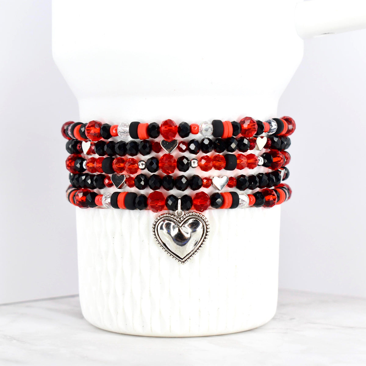 Stack of beaded bracelets in red, black, and silver. The bracelets feature a variety of beads, including faceted and round beads. Each bracelet has a silver heart charm. The bracelets are displayed on a white ceramic container.