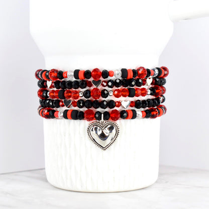 Stack of beaded bracelets in red, black, and silver. The bracelets feature a variety of beads, including faceted and round beads. Each bracelet has a silver heart charm. The bracelets are displayed on a white ceramic container.