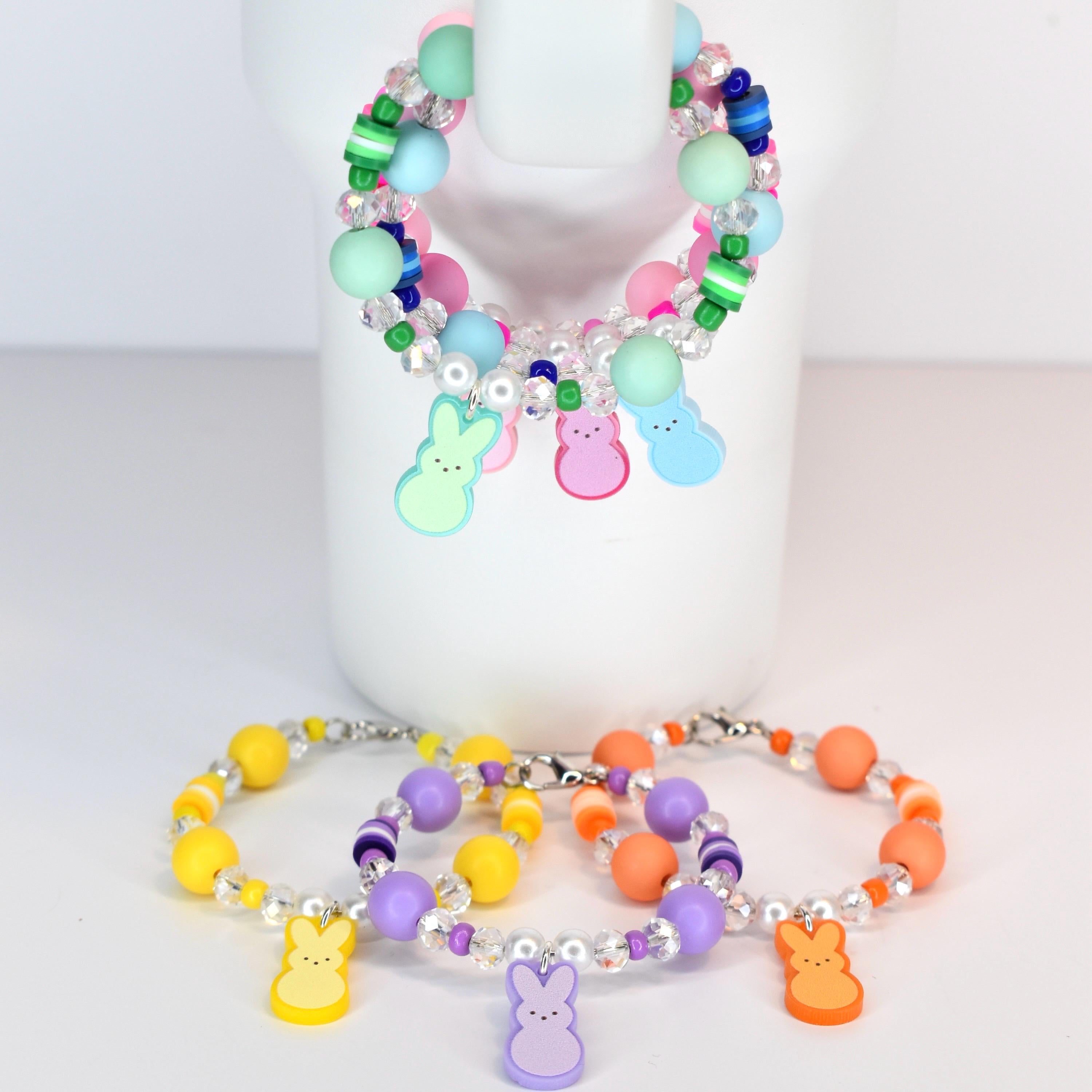 Colorful beaded bracelets with pastel and clear beads. The bracelets feature charms of Easter bunnies in yellow, orange, purple, and green. One bracelet is hanging on a white mug. The other bracelets are on a white surface.