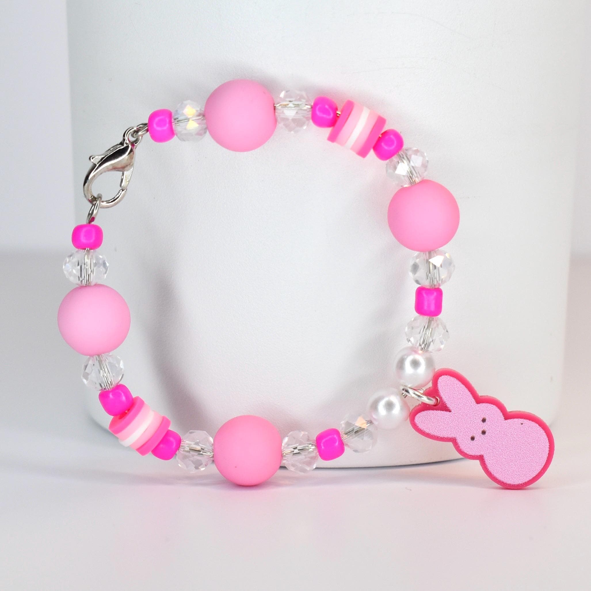 A pink beaded bracelet with a Peep charm. The bracelet features large pink beads, clear crystal beads, and small pink and white striped beads. A pink Peep-shaped charm hangs from the bracelet, which is secured with a silver clasp.