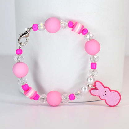A pink beaded bracelet with a Peep charm. The bracelet features large pink beads, clear crystal beads, and small pink and white striped beads. A pink Peep-shaped charm hangs from the bracelet, which is secured with a silver clasp.