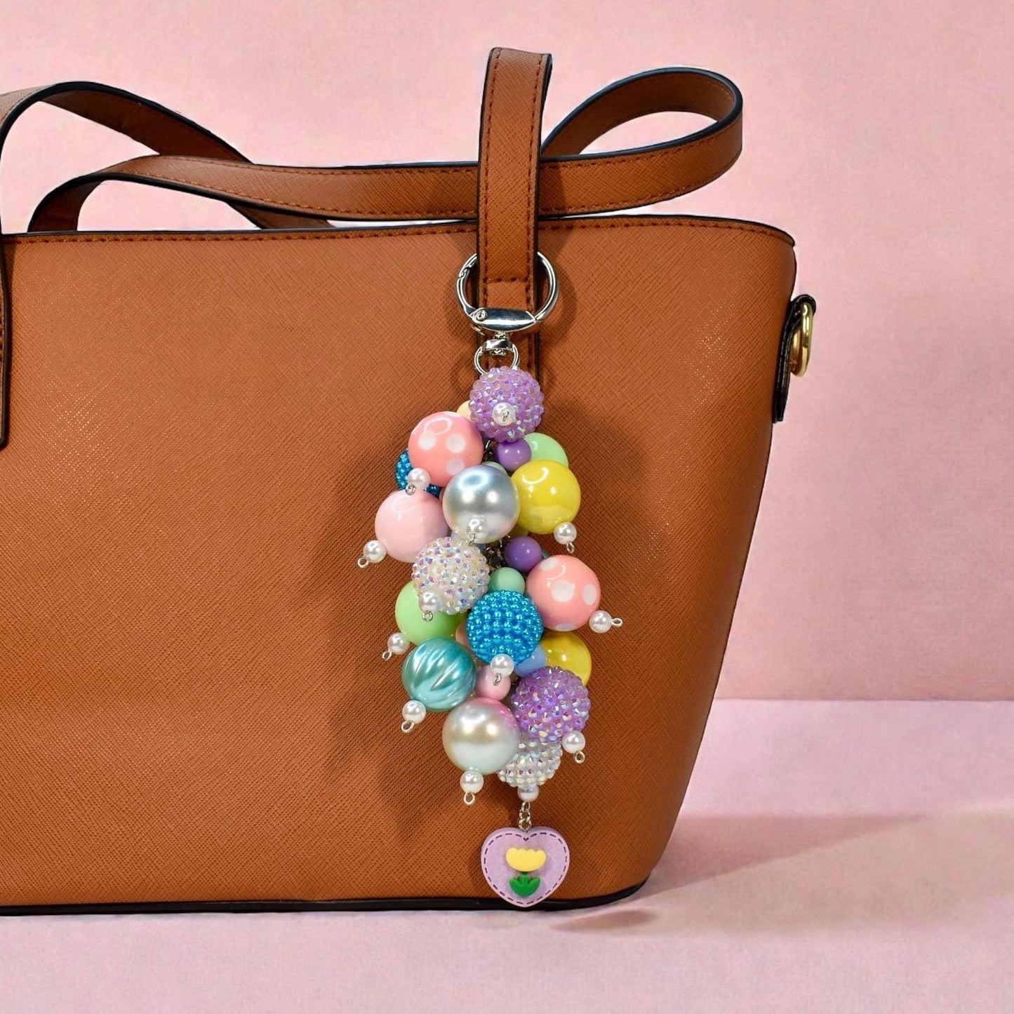 A colorful beaded keychain with a variety of pastel and iridescent beads, including pink, blue, yellow, and purple. The keychain is attached to a brown leather tote bag and features a heart-shaped charm with a flower design.