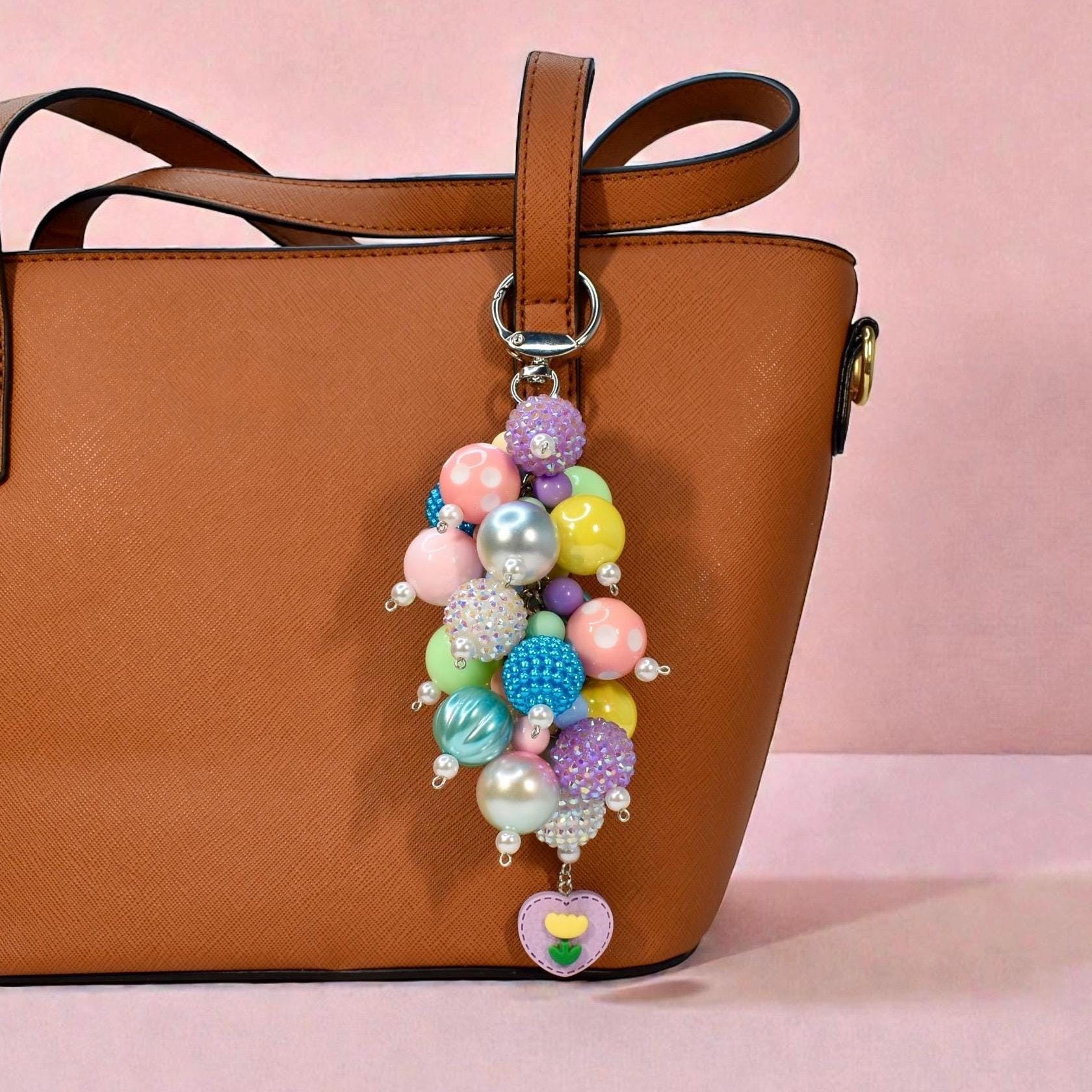 A colorful beaded keychain with a variety of pastel and iridescent beads, including pink, blue, yellow, and purple. The keychain is attached to a brown leather tote bag and features a heart-shaped charm with a flower design.
