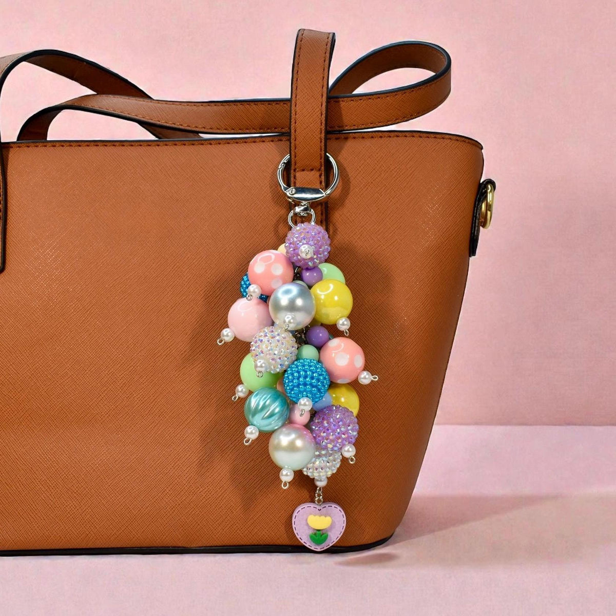 A colorful beaded keychain with a variety of pastel and iridescent beads, including pink, blue, yellow, and purple. The keychain is attached to a brown leather tote bag and features a heart-shaped charm with a flower design.