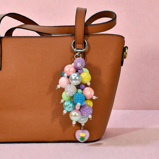 A colorful beaded keychain with a variety of pastel and iridescent beads, including pink, blue, yellow, and purple. The keychain is attached to a brown leather tote bag and features a heart-shaped charm with a flower design.
