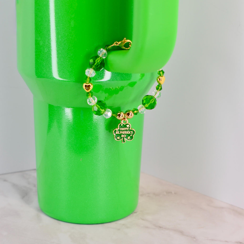 A St. Patrick's Day bracelet with green and clear beads, gold accents, and a shamrock charm that says "Happy St. Patrick's Day." The bracelet is displayed on a bright green tumbler.